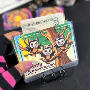 May include: A clear plastic card holder with a black metal binder clip, holding several cards and a stack of US five-dollar bills. The cards feature a cartoon illustration of three bats on a tree branch, with the text "Batty Summerween".
