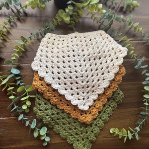 May include: A stack of three crocheted square cloths in cream, rust, and olive green. The cloths have a scalloped edge and a grid-like open weave. The items are arranged on a dark wood surface with greenery.