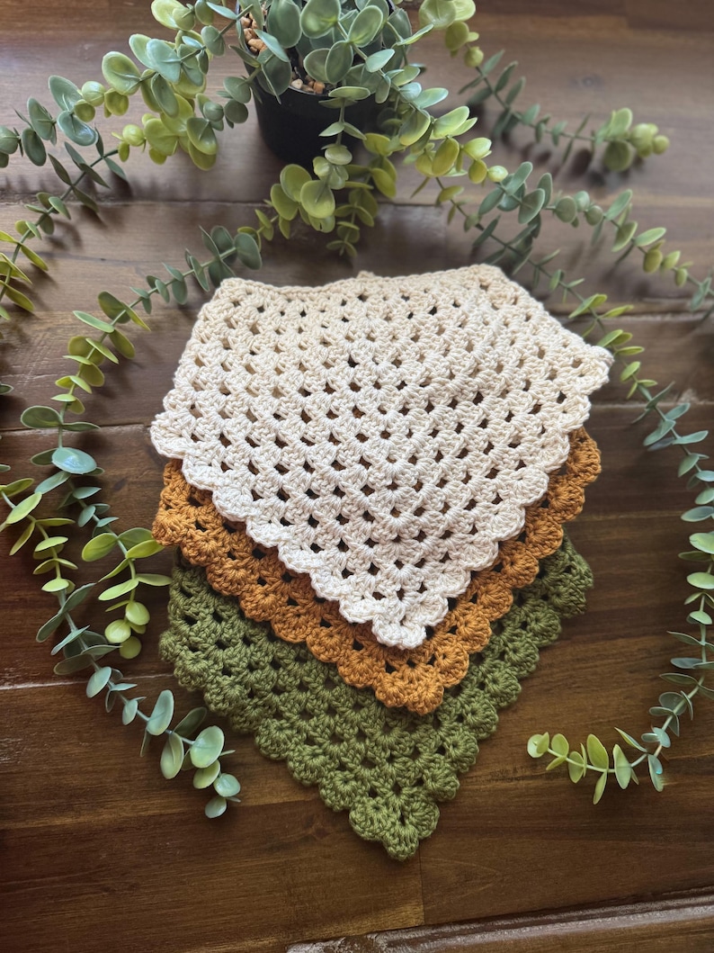 May include: A stack of three crocheted square cloths in cream, rust, and olive green. The cloths have a scalloped edge and a grid-like open weave. The items are arranged on a dark wood surface with greenery.