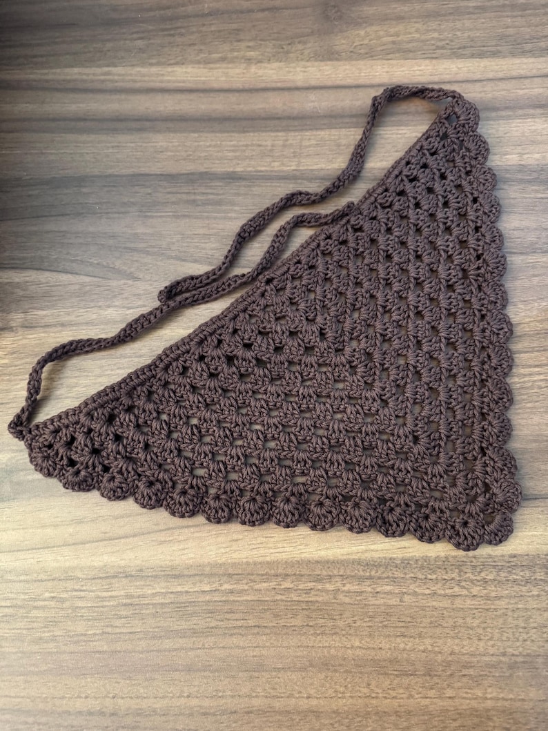 May include: A brown crochet bandana with a scalloped edge and long ties. The triangular scarf is made of a textured, open-weave fabric. The bandana is designed to be tied around the neck or head.
