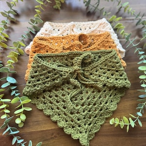 May include: Four handmade crochet bandanas in shades of green, orange, and beige are stacked on a wooden surface. The bandanas have a textured, open weave design. Greenery surrounds the bandanas, adding a natural touch to the display.