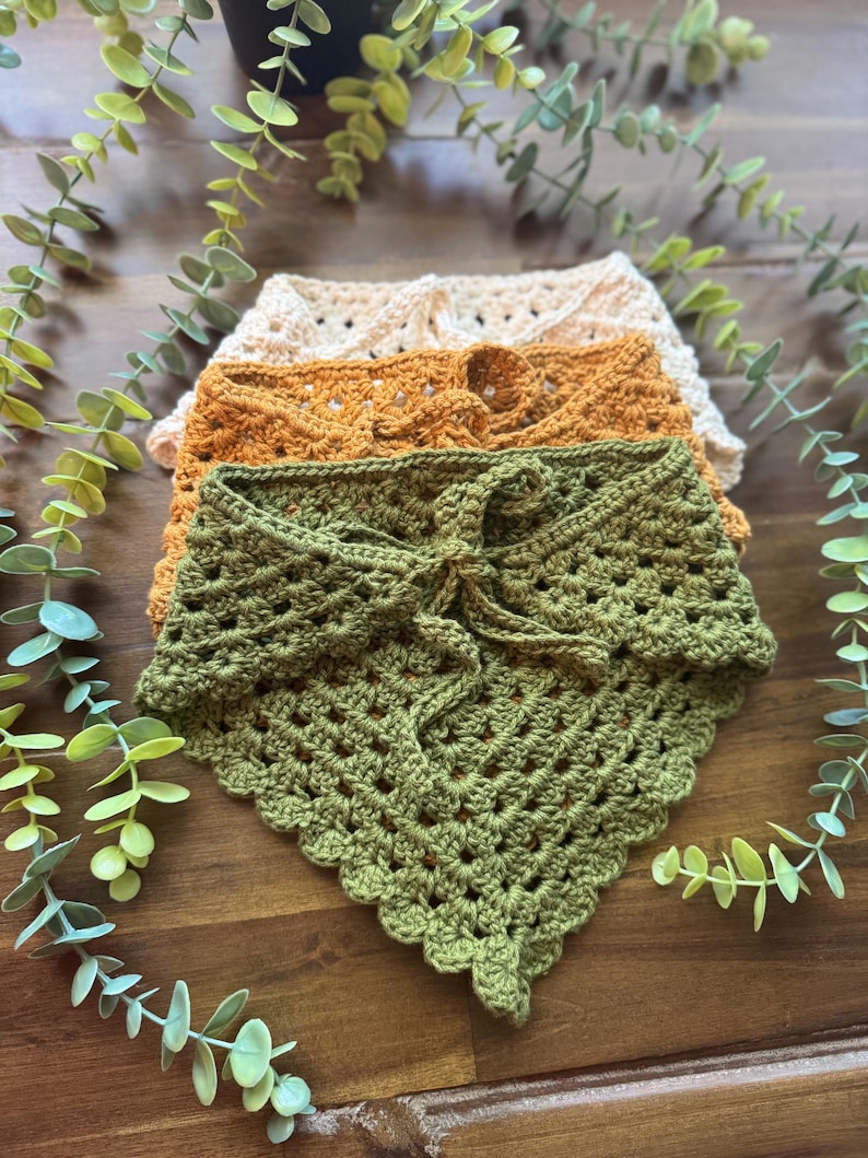 May include: Four handmade crochet bandanas in shades of green, orange, and beige are stacked on a wooden surface. The bandanas have a textured, open weave design. Greenery surrounds the bandanas, adding a natural touch to the display.