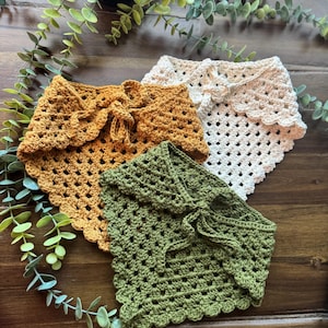 May include: Three crocheted shawls in shades of mustard yellow, cream, and olive green are displayed on a wooden surface. Each shawl features a scalloped edge and a tie closure at the front, showcasing a handmade aesthetic.