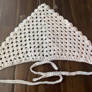 May include: Cream-colored, handmade crochet headscarf. The triangular scarf features an open, lacy pattern and a scalloped edge. It has long, crocheted ties for securing around the head. The scarf is displayed on a wooden surface.