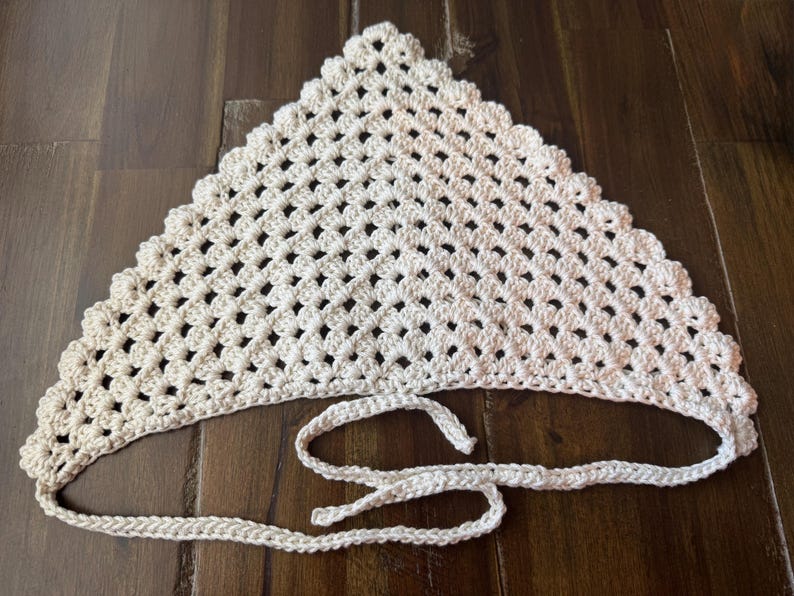 May include: Cream-colored, handmade crochet headscarf. The triangular scarf features an open, lacy pattern and a scalloped edge. It has long, crocheted ties for securing around the head. The scarf is displayed on a wooden surface.