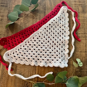 May include: Two handmade crochet bandanas. One is red, the other is white. Both have a triangular shape with scalloped edges and long ties. The bandanas are displayed on a wooden surface with green leaves.