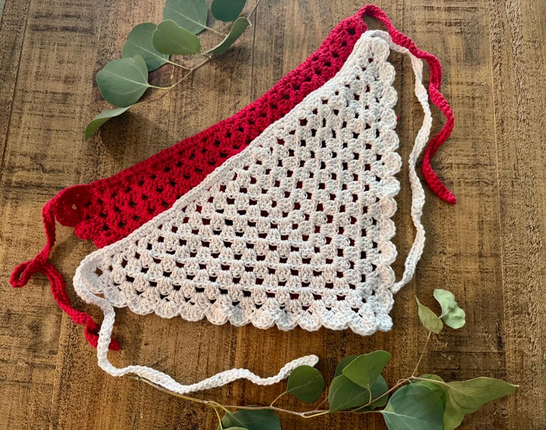 May include: Two handmade crochet bandanas. One is red, the other is white. Both have a triangular shape with scalloped edges and long ties. The bandanas are displayed on a wooden surface with green leaves.
