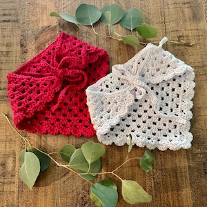 May include: Two crocheted boleros in red and white, each with a scalloped edge and tie closure. The openwork design and soft texture are visible. The boleros are displayed on a wooden surface with eucalyptus leaves.