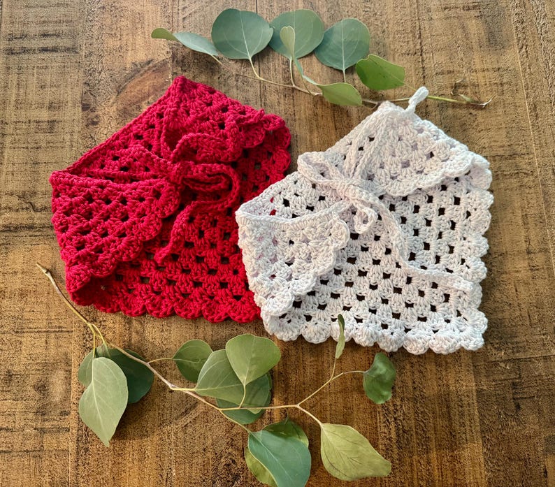 May include: Two crocheted boleros in red and white, each with a scalloped edge and tie closure. The openwork design and soft texture are visible. The boleros are displayed on a wooden surface with eucalyptus leaves.