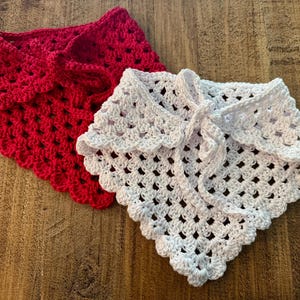 May include: Two handmade crochet shawls. One is a vibrant red, the other is white. Both have a scalloped edge and a tie closure. The open weave design is visible. These shawls are perfect for adding a touch of handmade charm to any outfit.