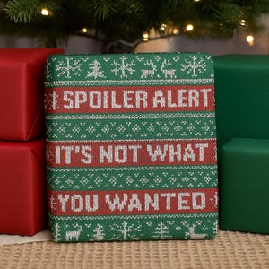 以下が含まれることがあります： 緑と赤のニット模様のギフトボックス。前面には「SPOILER ALERT」、「IT'S NOT WHAT」、「YOU WANTED」の文字が表示されています。 ギフトは他の包装されたギフトに囲まれています。