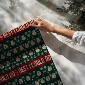 May include: A green and red Christmas-themed knitted blanket with the text "BEST I COULD DO" and snowman and snowflake patterns. The blanket is being held by a person.