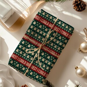 May include: A wrapped gift with green wrapping paper featuring a repeating pattern of snowmen and snowflakes. The words "BEST I COULD DO" are printed in red. The gift is tied with twine and is surrounded by Christmas ornaments.