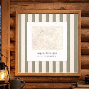 May include: Framed print of Aspen, Colorado, featuring a topographic map in neutral tones. The print is set against a striped background of white and muted green, encased in a wooden frame. Coordinates are displayed below the text.