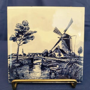May include: Square ceramic tile featuring a blue and white illustration of a Dutch windmill, a tree, and a waterway. The artwork is set against a cream-colored background. The tile is displayed on a gold-colored metal stand.