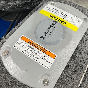 May include: A gray warning sign with black text that reads "AVOID SERIOUS OR FATAL INJURY. DO NOT OCCUPY SEAT WHEN SPEED EXCEEDS 5 MPH." The sign is attached to a gray surface with a black Lund Boats logo and the word "CAUTION" in yellow.
