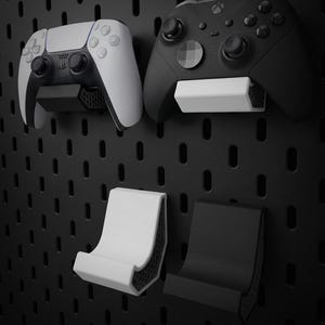 Puede incluir: Un panel perforado negro con dos mandos de juego y dos soportes para mandos. Un mando blanco y negro está montado en un soporte blanco. Un mando negro está montado en un soporte negro. Se muestran dos soportes adicionales.