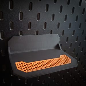 Könnte beinhalten: Ein schwarzes Lochwandregal mit einem orangefarbenen Wabenmuster. Das Regal ist für die Befestigung an einem Lochwandsystem konzipiert. Das Regal besteht aus einem strapazierfähigen Material und ist für kleine Gegenstände konzipiert.