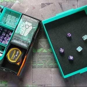 Op de afbeelding: Een turquoise organizer met vakken voor dobbelstenen, spelkaarten en een meetlint. De organizer is open en de vakken zijn gevuld met verschillende dobbelstenen. De organizer staat op een grijs en groen gepatroneerd oppervlak.