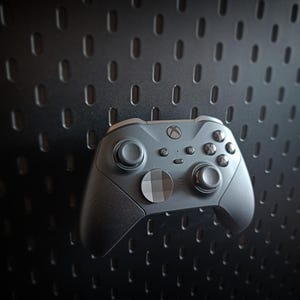 Puede incluir: Un mando de Xbox gris con detalles en negro, que incluye un botón circular central y varios botones de control. El mando se encuentra sobre un fondo oscuro y texturizado con un patrón de pequeños agujeros espaciados.