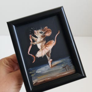 Peut inclure: Une peinture à l'huile dans un cadre noir représentant une souris brune portant un tutu rose clair et tenant un ruban rose. La souris est posée sur une patte sur un fond sombre, avec les initiales TA de l'artiste visibles en bas à droite.