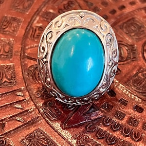 May include: A silver ring with an oval, bright turquoise stone. The ring has an ornate, swirling design around the stone. The ring is set on a textured, reddish-brown surface.