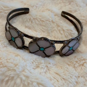 May include: Silver-toned cuff bracelet featuring three flower-shaped accents. Each flower has a light pink petal design with a turquoise center. The bracelet is set against a soft, cream-colored background.