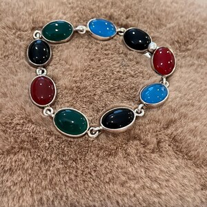 May include: A silver bracelet featuring oval-shaped gemstones in various colors, including green, blue, black, and red. The gemstones are set in a silver link chain. The bracelet is displayed on a brown, textured surface.