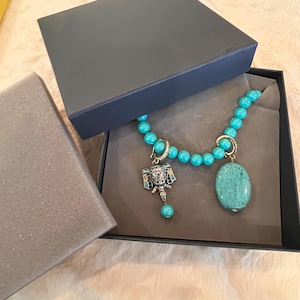 May include: A turquoise beaded necklace with a pendant and a large oval stone, presented in a dark gray gift box. The necklace features a detailed pendant with silver and turquoise accents. The box is open, revealing the jewelry on a gray fabric.