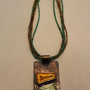 May include: A copper-toned pendant necklace with a rectangular pendant featuring two inset, irregularly shaped stones in shades of yellow and green. The necklace has multiple strands of brown and green beads.