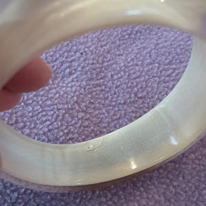 K&ouml;nnte beinhalten: Ein breiter, durchscheinender, cremewei&szlig;er Armreif mit einer glatten, polierten Oberfl&auml;che. Das kreisf&ouml;rmige Accessoire weist einen subtilen, perlmuttartigen Glanz im gesamten Material sowie eine kleine, deutliche Vertiefung auf der Innenseite auf.