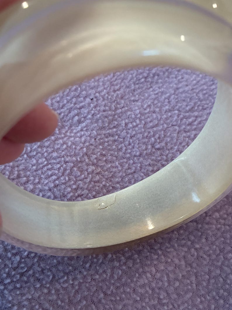 K&ouml;nnte beinhalten: Ein breiter, durchscheinender, cremewei&szlig;er Armreif mit einer glatten, polierten Oberfl&auml;che. Das kreisf&ouml;rmige Accessoire weist einen subtilen, perlmuttartigen Glanz im gesamten Material sowie eine kleine, deutliche Vertiefung auf der Innenseite auf.