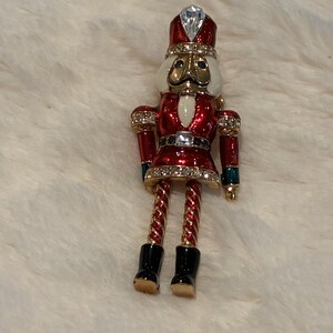 May include: A nutcracker brooch featuring red enamel, gold-tone metal, and clear crystal accents. The figure wears a tall hat with a clear stone, a red jacket with a white chest piece, and black boots. The arms and legs are articulated.