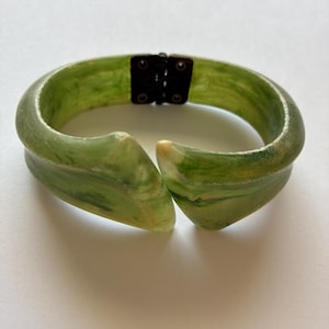 May include: Green and white marbled bangle bracelet with a hinged clasp. The bracelet has a rounded shape with a slight point on each end. The clasp is a dark metal. The bracelet is made of a translucent material.