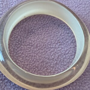 K&ouml;nnte beinhalten: Ein kreisf&ouml;rmiger Armreif mit einer klaren, transparenten Au&szlig;enschicht und einem glatten, undurchsichtigen wei&szlig;en Innenband. Das Design ist schlicht und einheitlich, mit einem abgerundeten Profil, das ein sauberes, geschichtetes Erscheinungsbild erzeugt.