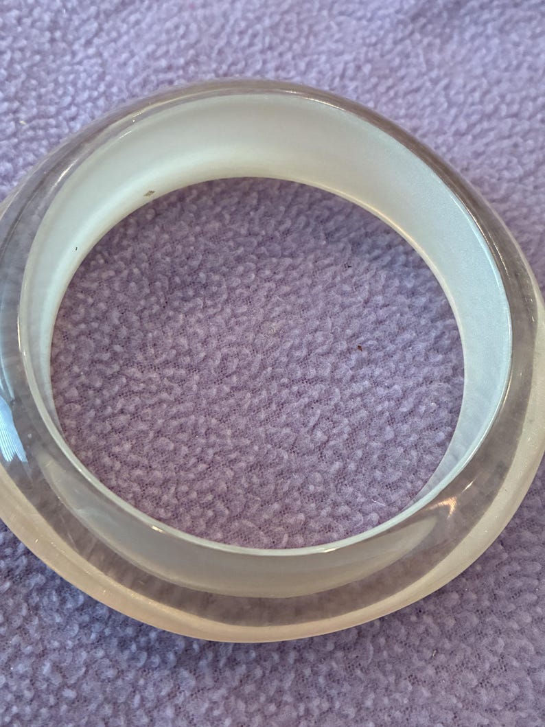 K&ouml;nnte beinhalten: Ein kreisf&ouml;rmiger Armreif mit einer klaren, transparenten Au&szlig;enschicht und einem glatten, undurchsichtigen wei&szlig;en Innenband. Das Design ist schlicht und einheitlich, mit einem abgerundeten Profil, das ein sauberes, geschichtetes Erscheinungsbild erzeugt.
