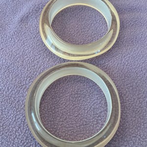 K&ouml;nnte beinhalten: Zwei kreisf&ouml;rmige Armreifen aus klarem, transparentem Material. Jeder Armreif verf&uuml;gt &uuml;ber eine glatte, abgerundete Au&szlig;enfl&auml;che und eine hohle Mitte, was ein schlichtes, unverziertes Design mit einer gl&auml;nzenden Oberfl&auml;che zur Geltung bringt.