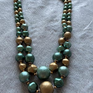 Könnte beinhalten: Eine zweireihige Halskette mit abwechselnd runden und facettierten Perlen in Metallic-Grün und mattem Gold. Die Perlen sind mit kleinen goldfarbenen Zwischenelementen aufgereiht und die Kette verfügt über einen Metallverschluss am oberen Ende.