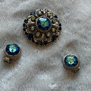 Könnte beinhalten: Ein Schmuckset bestehend aus einer runden Brosche und einem Paar passender Clip-Ohrringe. Jedes Stück verfügt über einen zentralen, schillernden blau-grünen Kristall, umgeben von kleineren blauen und hellgrünen Steinen in einer gedrehten silberfarbenen Metallfassung.
