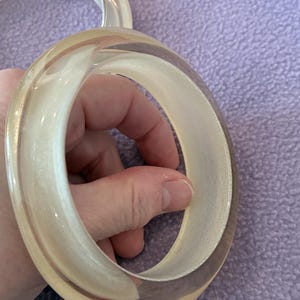 K&ouml;nnte beinhalten: Zwei kreisf&ouml;rmige Armreifen aus transparentem Harz. Jeder Armreif hat eine glatte, abgerundete Au&szlig;enfl&auml;che und einen massiven, undurchsichtigen cremewei&szlig;en Innenkern, der ein geschichtetes, dreidimensionales Erscheinungsbild im klaren Material erzeugt.