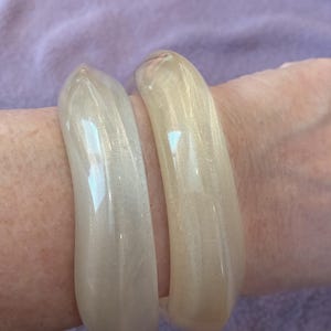 K&ouml;nnte beinhalten: Zwei glatte, durchscheinende Armreifen in Off-White mit einem dezenten Perlmuttschimmer. Die Armreifen haben eine abgerundete, polierte Oberfl&auml;che und eine durchgehende Kreisform, die das Licht auf ihrem gl&auml;nzenden Finish reflektiert.