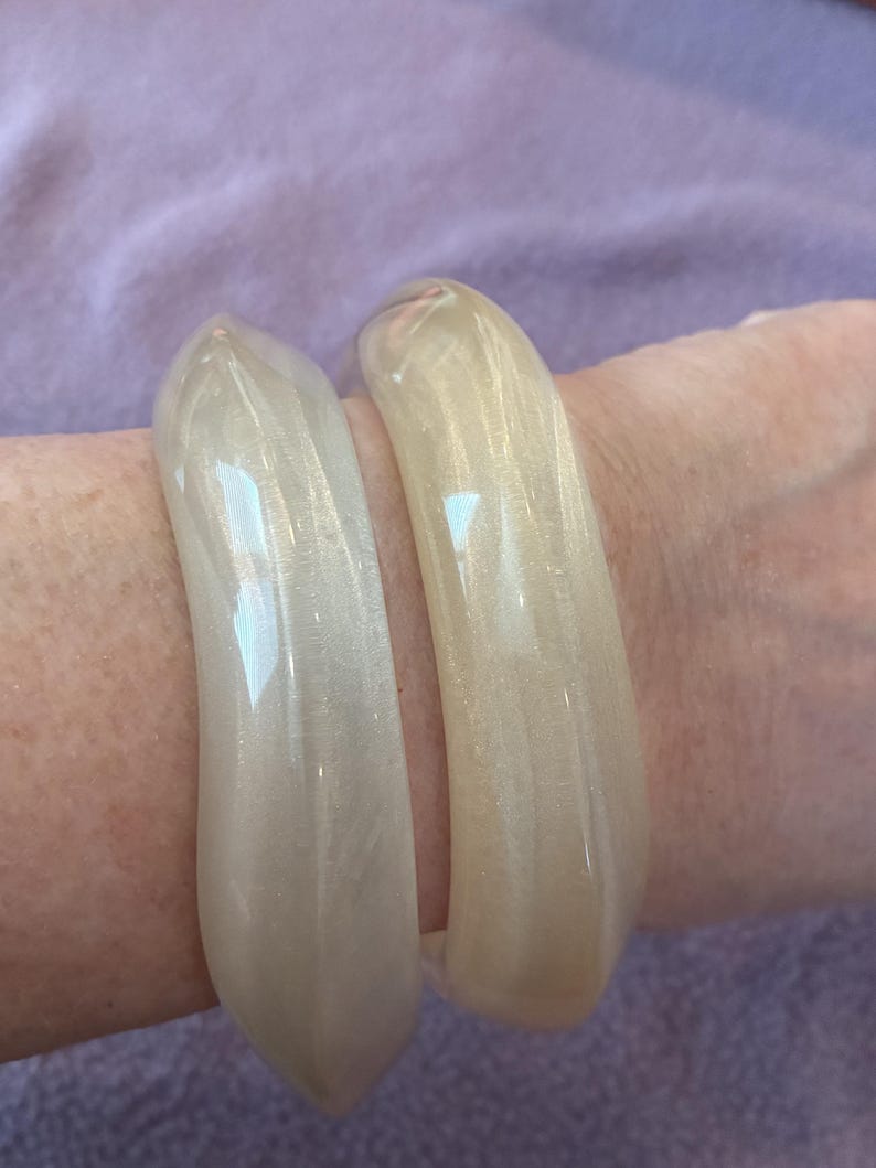 K&ouml;nnte beinhalten: Zwei glatte, durchscheinende Armreifen in Off-White mit einem dezenten Perlmuttschimmer. Die Armreifen haben eine abgerundete, polierte Oberfl&auml;che und eine durchgehende Kreisform, die das Licht auf ihrem gl&auml;nzenden Finish reflektiert.