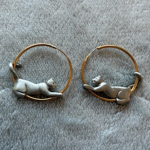 Könnte beinhalten: Ein Paar Creolen mit einem dünnen, goldfarbenen, kreisförmigen Drahtrahmen. Jeder Ohrring enthält eine kleine, silberfarbene Katzenfigur, die am Ring befestigt ist und in einer liegenden oder sich streckenden Pose am unteren Bogen dargestellt ist.