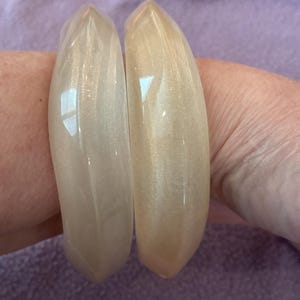 K&ouml;nnte beinhalten: Zwei durchscheinende, cremewei&szlig;e Armreifen mit einer glatten, polierten Oberfl&auml;che und einem dezenten Perlmuttschimmer. Die Armreifen haben eine leicht facettierte, geometrische Au&szlig;enkante und ein abgerundetes Innenband mit Reflexionen.