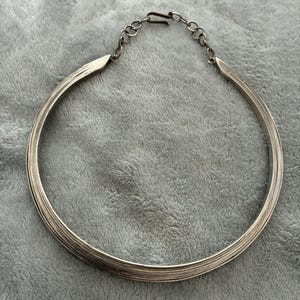 May include: A silver-toned metal collar necklace featuring a rigid, semi-circular band with a textured, multi-line finish. The necklace includes a short chain extension on each side, connected by a simple hook clasp at the back for closure.