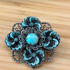 Könnte beinhalten: Florale Brosche mit einem türkis-schwarzen Blumendesign. Die Brosche hat vier türkisfarbene Blumen mit schwarzen Details, einen zentralen türkisfarbenen Stein und einen silbernen Filigranrand. Die Brosche ist vor einem Holzgrund platziert.