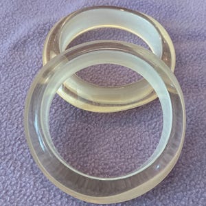 K&ouml;nnte beinhalten: Zwei kreisf&ouml;rmige Armreifen aus klarem, durchscheinendem Material. Die Armb&auml;nder haben eine glatte, abgerundete Oberfl&auml;che mit poliertem Finish, was eine einfache und klare &Auml;sthetik erzeugt. Das klare Material l&auml;sst Licht hindurch und betont die kreisf&ouml;rmige Form sowie die gleichm&auml;&szlig;ige Dicke der B&auml;nder.