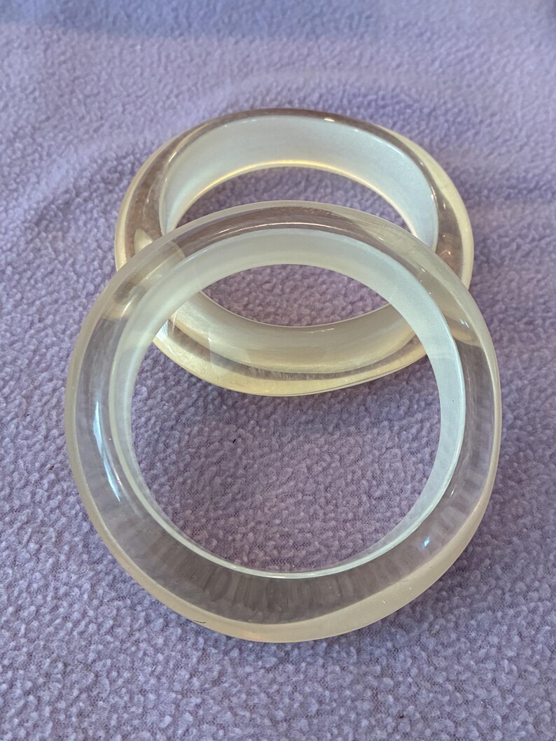 K&ouml;nnte beinhalten: Zwei kreisf&ouml;rmige Armreifen aus klarem, durchscheinendem Material. Die Armb&auml;nder haben eine glatte, abgerundete Oberfl&auml;che mit poliertem Finish, was eine einfache und klare &Auml;sthetik erzeugt. Das klare Material l&auml;sst Licht hindurch und betont die kreisf&ouml;rmige Form sowie die gleichm&auml;&szlig;ige Dicke der B&auml;nder.
