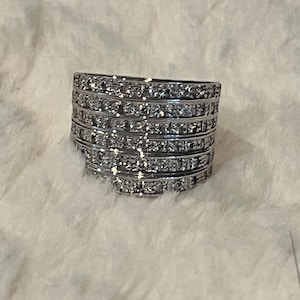 May include: A silver-toned ring with multiple bands, each encrusted with small, sparkling clear stones. The ring has a layered design, creating a wide, textured appearance. The stones catch the light, giving the ring a brilliant shine.