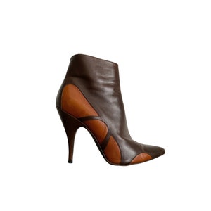 May include: A brown and tan leather ankle boot with a pointed toe and a high stiletto heel. The boot features a unique design with cut-out details and a sleek, modern silhouette. The boot is set against a white background.